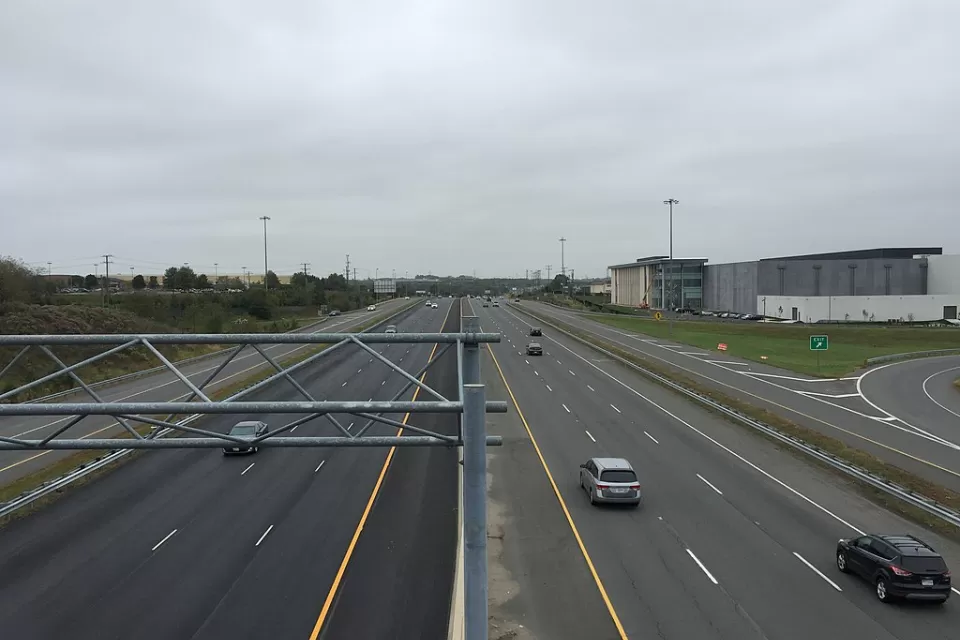 A wide highway with a few cars on a gloomy day. Low-rise commercial buildings and more car infrastructure is visible to the side of the road.